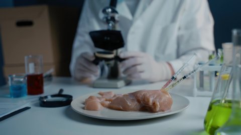 Food quality control scientist works with microscope in laboratory. Syringes with antibiotics in raw meat, injections with genetically modified organisms in chicken, drugs and chemicals in food.