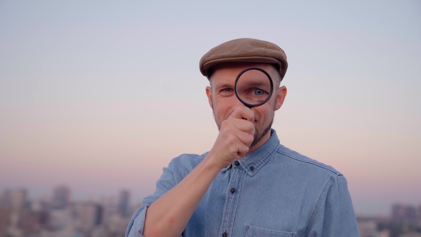 Attractive man looking with magnifying glass at camera wearing jeans shirt and peaked cap. Male searching sales or discounts at sunset using loupe with urban view. Shopping concept. High quality 4k Royalty-Free Stock Footage #1088945327