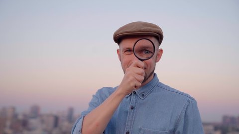 Attractive man looking with magnifying glass at camera wearing jeans shirt and peaked cap. Male searching sales or discounts at sunset using loupe with urban view. Shopping concept. High quality 4k
