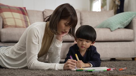 slow motion of loving taiwanese mom lying prone on floor teaching her preschool son to use color pencils drawing on paper at home