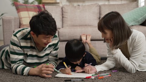 cheerful chinese family of three having fun lying prone on living room floor drawing picture together. the mother laughs at her baby son’s silly talks