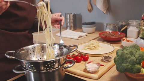 Cropped shot of woman in apron taking freshly cooked homemade pasta from colander and serving it on plate while making dinner in kitchen at home