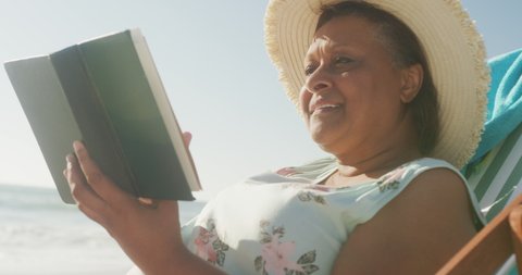 Smiling senior african american woman lying on sunbed and reading book on sunny beach. healthy, active retirement beach holiday.