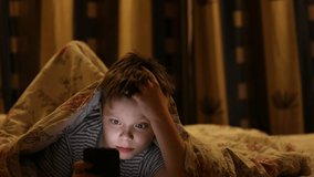 the boy lies under the blanket and plays games on the smartphone, at night