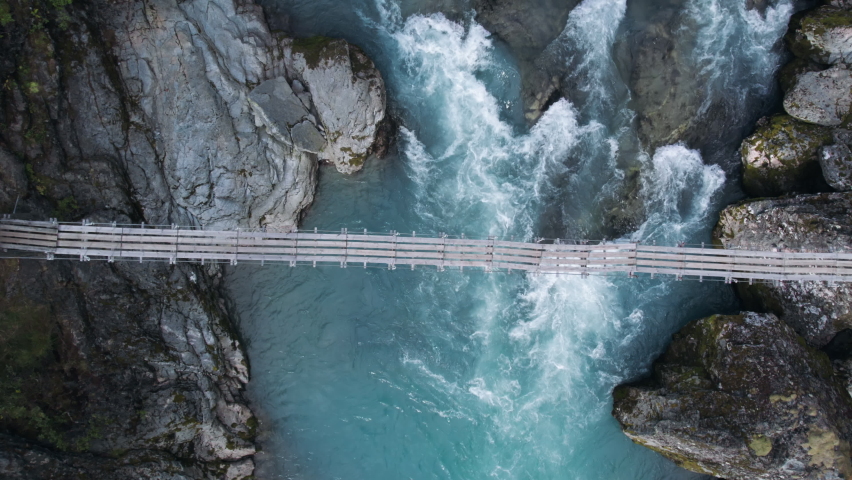Incredible and adventure inspiring aerial drone shot of adrenaline seeking and exploring young hiker woman walk over old epic wooden suspension bridge in scandinavian forest. Amazing light blue