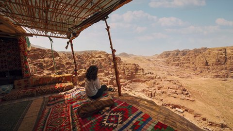 Cinemagraph seamless video loop of a young female woman in Petra, Jordan, sitting in a bedouin hut with colorful vibrant carpets and a view of the historic roman theater carved into sandstone rock.