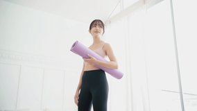 Smiling Asian woman wearing sports bra and yoga pants holding a rolled-up yoga mat standing in a bedroom with sunlight from the window. Slow-motion portrait video.