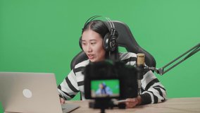 A Smiling Asian Woman Gamer With Headphone And Computer Playing Game On Mobile Phone While Shooting Video By Camera On The Green Screen Background
