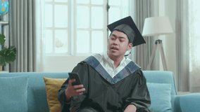 Excited Asian Man Showing Off A University Certificate To The Family During An Online Video Call By Mobile Phone. Male Graduate Wearing A Graduation Gown And Cap On The Living Room
