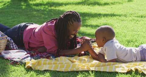 Video of happy african american father and son having picnic on grass, arm wrestling. family, togetherness, spending quality time together outdoors.