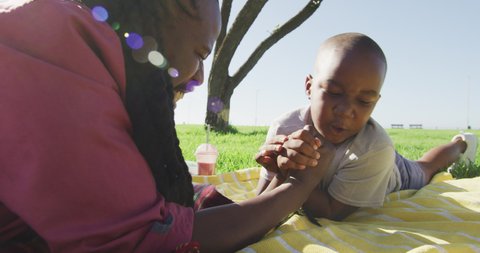 Video of happy african american father and son having picnic on grass, arm wrestling. family, togetherness, spending quality time together outdoors.