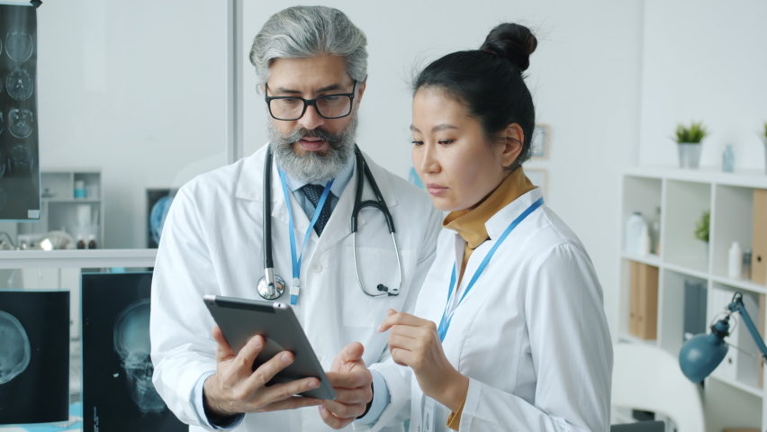 Male and female doctors looking at tablet screen talking and smiling indoors in modern clinic. Professional communication and technology concept. Royalty-Free Stock Footage #1088959069