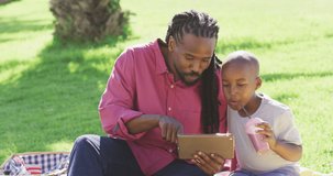 Video of happy african american father and son having picnic outdoors and using tablet. family, togetherness, spending quality time together outdoors.