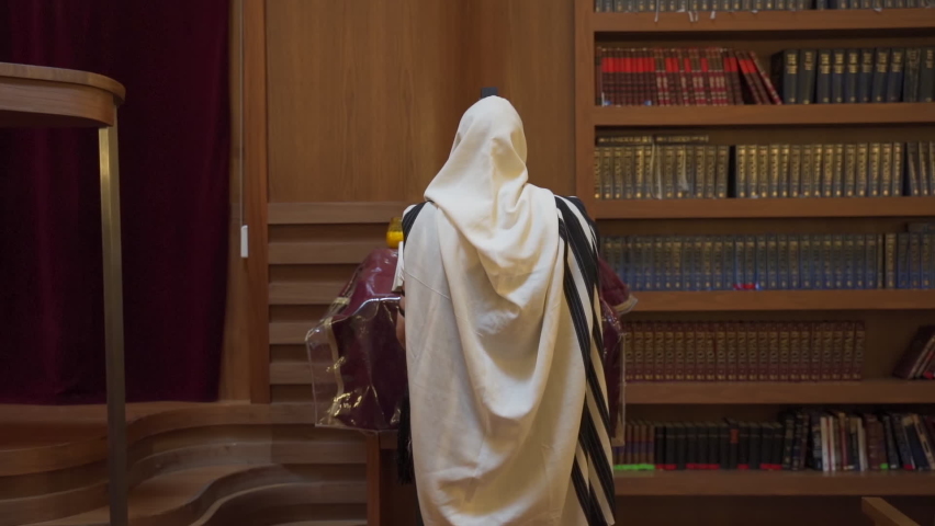 A Jew prays in the synagogue. A man stands near the presidium and reads a book. Rearview
