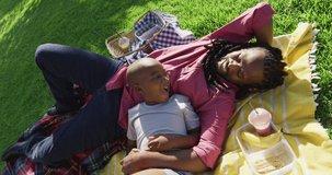 Video of happy african american father and son having picnic on grass and resting on blanket. family, togetherness, spending quality time together outdoors.