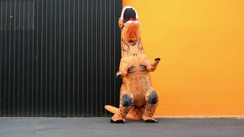 One happy and funny dinosaur costume dancing in the street with a orange colorful background - t-rex having fun - funny man inside of a costume of dino
 | Shutterstock HD Video #1088959675
