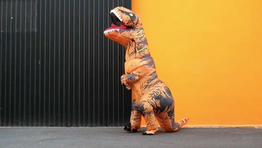 One happy and funny dinosaur costume dancing in the street with a orange colorful background - t-rex having fun - funny man inside of a costume of dino
 | Shutterstock HD Video #1088959675