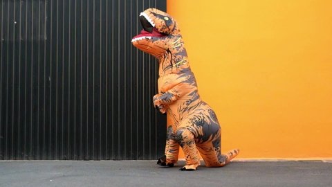 One happy and funny dinosaur costume dancing in the street with a orange colorful background - t-rex having fun - funny man inside of a costume of dino
