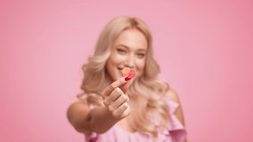 Attractive Blonde Female Showing And Eating Heart Shaped Jelly Candy Smiling To Camera Enjoying Taste Posing Standing Over Pink Studio Background. Sweet Tooth, Sugary Food Concept