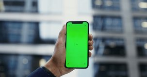 A man's hand in a business suit holds a mobile phone with a green screen on the background of a modern building. Copy space, mockup, chroma key. Video in 4k, red komodo