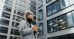 Young stylish African-American businesswoman in a medical mask on her face is typing on a mobile phone while standing near a modern office building. Video in 4k, red komodo