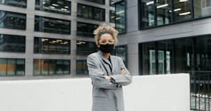 African-American businesswoman wearing a medical mask on her face looks at the camera while standing near the office building with her arms crossed over her chest. Video in 4k, red komodo