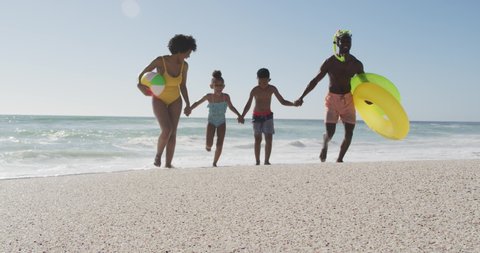 Smiling african american family with inflatables running on sunny beach. healthy, active family beach holiday.