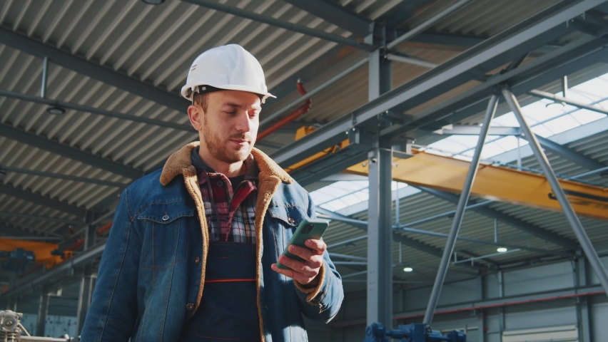 Portrait smiling engineer man with helmet and uniform use mobile phone walking in factory. Communication, worker. Royalty-Free Stock Footage #1088966087