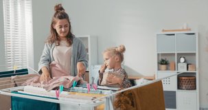 Woman performs household chores around the house, hangs clean clothes on dryer in laundry room, child finds fun, holds basket of clips, spends time with mom.