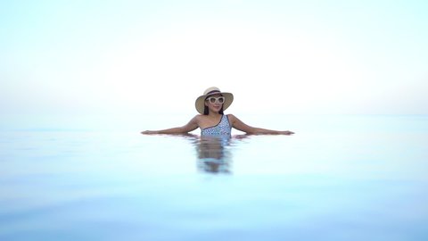 Beautiful woman standing in calm swimming pool with large hat and sunglasses relaxing in sunshine. Glamor and beauty concept with exclusive luxury tropical resort destination. Reflection in water.