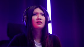 Gamer losing moment. Young asian pretty woman sitting on chair with computer pc. Happy female Professional Streamer chinese wearing headphone playing game online in dark room neon light.