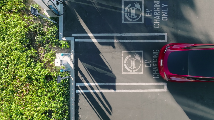 Vertical view on red electric vehicle backing up to park at the EV charging only station. Top down aerial view on zero emission modern vehicle parking to recharge battery in urban landscape at sunset