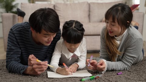 closeup view of happy asian father. mother and little girl lying prone on floor enjoying drawing picture together at home. family love and happy childhood concept