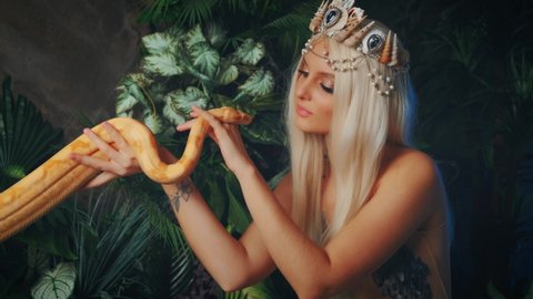 myth creature Nagga woman touches white milk Boa constrictor albino snake. Fantasy asp girl, blonde long hair, looks at serpent. Silver costume body tail of Lady viper. Backdrop green tropical palants
