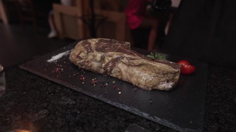 The chef places a large raw tomahawk steak on a grill rack built into the table at the restaurant. An exquisite piece of aged meat. Beef barbecue. Hands in black gloves.