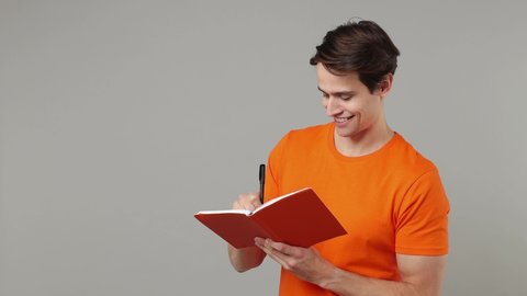 Happy charismatic blithesome fancy young brunet man student 20s years old wears orange t-shirt writing down in exercise book notebook diary notes isolated on plain grey wall background studio portrait