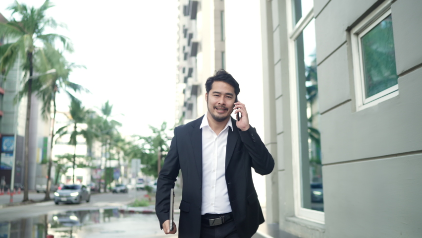 Asian Businessman Smiling Wearing a Black Suit is Rush Running On the Streets Of the City to Work. in Hand holding a Laptop using Smartphone. Urban lifeStyle Concept.
 Royalty-Free Stock Footage #1088984451