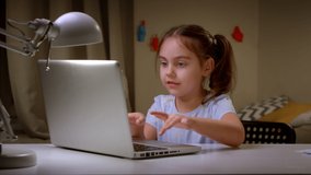 Preschooler girl watches a lesson online and studies at home, writes down a synopsis. Cute little girl takes notes while looking computer screen, following a professor doing lessons on a video call.