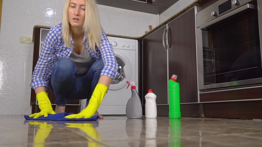 young Caucasian woman 25-35 years old washes floor while squatting on her knees. She runs blue rag across floor, collecting debris and dust. Yellow gloves. There are cleaning products in background.