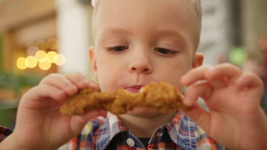 Portrait hungry blond 4-year-old boy sitting fast food and eating fried chicken in breadcrumbs. Caucasian kid satisfies his hunger eating deep-fried chicken from fast food. Junk food from fast food. | Shutterstock HD Video #1088990699