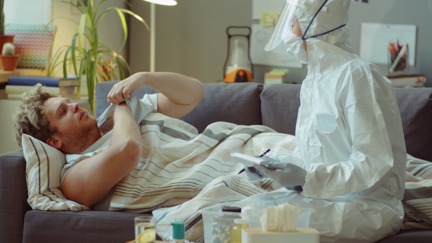 Doctor in protective uniform taking temperature of coronavirus patient, writing prescription and advising him medications during at-home checkup