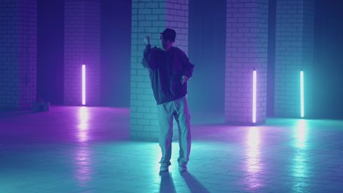 A steel man dances hip-hop freestyle in a modern style in a hall with neon light in purple blue colors. Male Professional Hip Hop Dancer