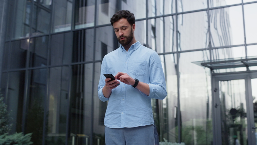 Attractive man stand texting at lively downtown area. Corporate people passing. Handsome bearded startup businessman thoughtfully typing message on phone. Focused manager using cellphone at office. Royalty-Free Stock Footage #1088993293