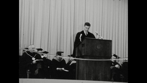 1960s: Man stands at podium with microphones, gives address, gestures. People sit on stage in caps and gowns, listen. Man looks restless, crosses legs, stretches hand.
