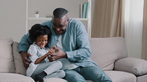 Happy african american family adult funny father playing with daughter sitting on sofa in living room dad tickling hugging cute kid girl enjoying playtime laughing have fun spending free time together