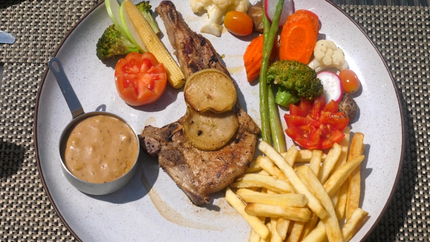 Woman hands with fork and knife cut a fresh roasted meat on a plate with vegetables in outdoor restaurant. A balanced diet. Pork chop with fries in a cafe. Top view. Slow motion