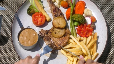 Woman hands with fork and knife cut a fresh roasted meat on a plate with vegetables in outdoor restaurant. A balanced diet. Pork chop with fries in a cafe. Top view. Slow motion