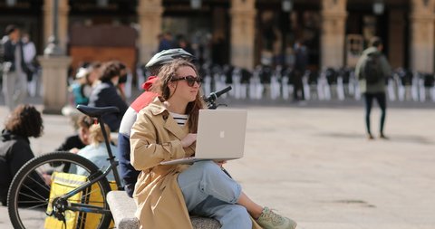 Salamanca, Spain- March 2022: Young girl uses a laptop sitting outdoors. Beautiful Young curly Woman with laptop having online work or education while sitting on the stairs in the middle of the city