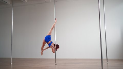 Athletic woman pole dancer in a blue suit performs acrobatic stunts with stretching elements on a pole in a light gym.