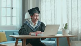 Happy Asian Man Wearing A Graduation Gown And Cap Use Laptop Computer In Living Room
